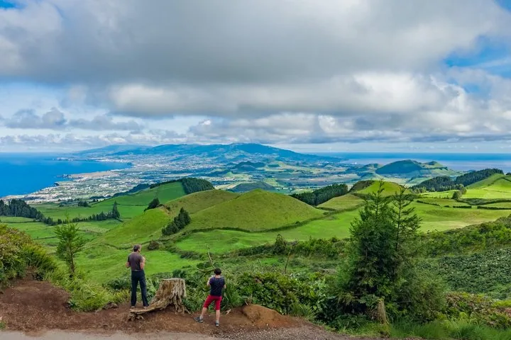 Two people standing on a dirt path overlooking green rolling hills.
